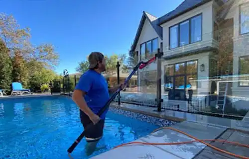 Man cleaning a luxurious backyard pool with a long-handled vacuum cleaner. The large house has modern windows and stone accents, with lush greenery in the background under a bright blue sky.