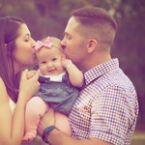 A happy family portrait: mother and father kissing their smiling baby girl, who is wearing a pink bow. The family is outdoors, bathed in soft, warm light.