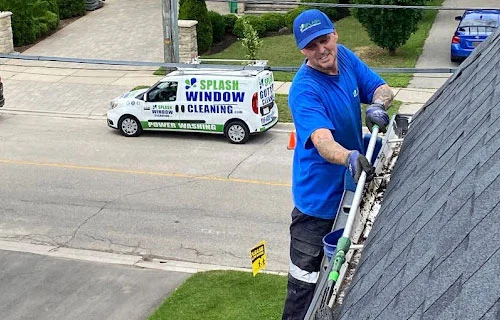 Man cleaning gutters on a house roof with a tool, wearing a blue shirt and hat. A "Splash Window Cleaning" van is parked on the street in the background. Gutter cleaning services.