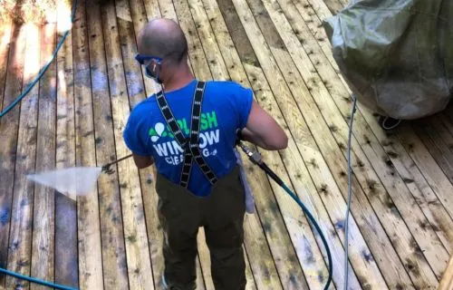 Man power washing a wooden deck. He wears safety glasses, waders, and a blue t-shirt with "Polish Window Cleaning" on the back. A high-pressure stream cleans the wood.