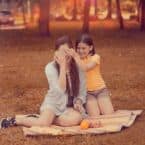 A young girl playfully covers her mother's eyes during a picnic in a sun-dappled park. The mother sits on a blanket, smiling, as her daughter kneels behind her in a yellow shirt and denim shorts.