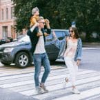 Family strolls across a crosswalk: Dad carries child on shoulders, mom walks beside, all smiling. City buildings and a dark SUV in the background.